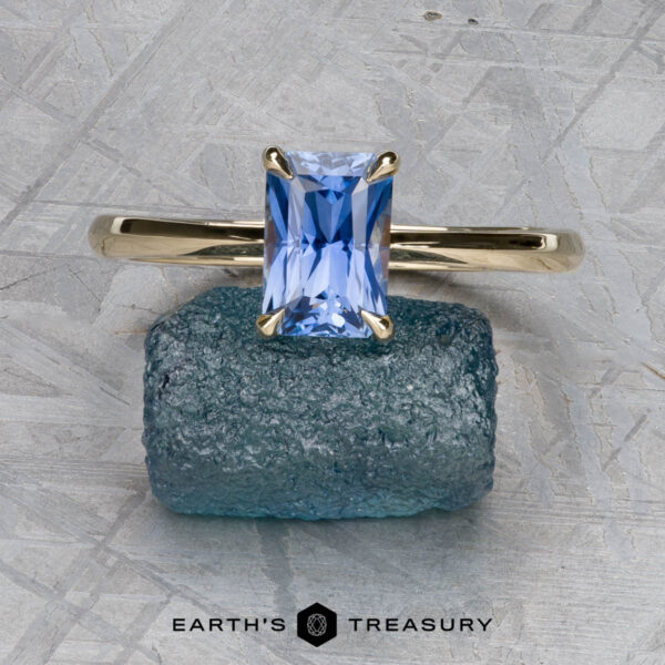 The Cathedral "Lyra" ring in 14K yellow gold with 1.34-carat Ceylon sapphire The Cathedral "Lyra" ring in 14K yellow gold with a 1.34-carat Ceylon sapphire is displayed upright on rough blue mineral, set against a textured gray background with "Earth’s Treasury" below.
