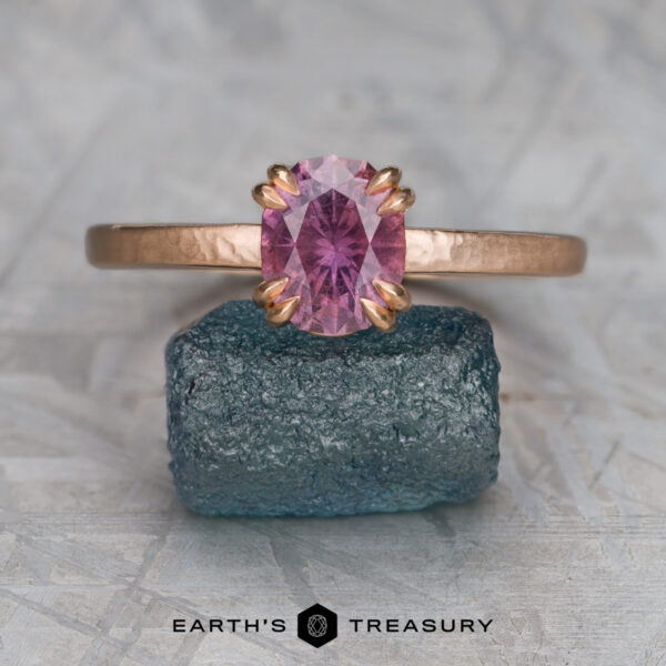 The “Capella” ring in 20K pink gold, hammered and brushed, with 1.63-Carat Umba Sapphire The “Capella” ring in 20K pink gold, hammered and brushed, with a 1.63-carat Umba Sapphire, stands upright on a rough blue-green stone. A light gray background and "EARTH'S TREASURY" text appear below.