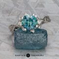 The "Juniper" ring in platinum, featuring a 1.63-carat Montana sapphire and accent diamonds, rests on a rough blue crystal against a silvery textured background. The "EARTH'S TREASURY" logo appears at the bottom.