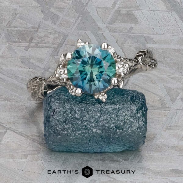 The "Juniper" ring in platinum with 1.63-carat Montana sapphire The "Juniper" ring in platinum, featuring a 1.63-carat Montana sapphire and accent diamonds, rests on a rough blue crystal against a silvery textured background. The "EARTH'S TREASURY" logo appears at the bottom.