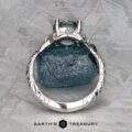 The "Juniper" ring in platinum with a 1.63-carat Montana sapphire stands upright on a rough blue stone, showcasing detailed band design. Set against a textured gray background. Text reads "Earth's Treasury.
