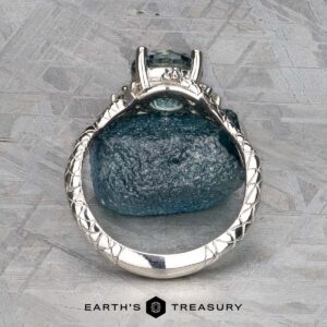 The "Juniper" ring in platinum with a 1.63-carat Montana sapphire stands upright on a rough blue stone, showcasing detailed band design. Set against a textured gray background. Text reads "Earth's Treasury.