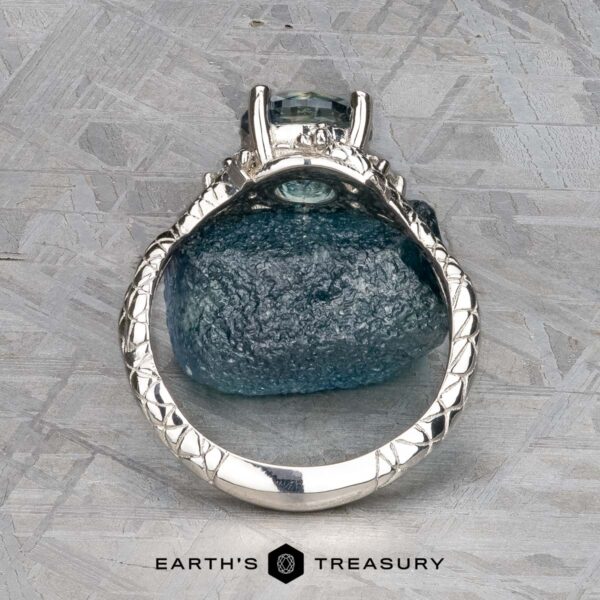 The "Juniper" ring in platinum with 1.63-carat Montana sapphire The "Juniper" ring in platinum with a 1.63-carat Montana sapphire stands upright on a rough blue stone, showcasing detailed band design. Set against a textured gray background. Text reads "Earth's Treasury.
