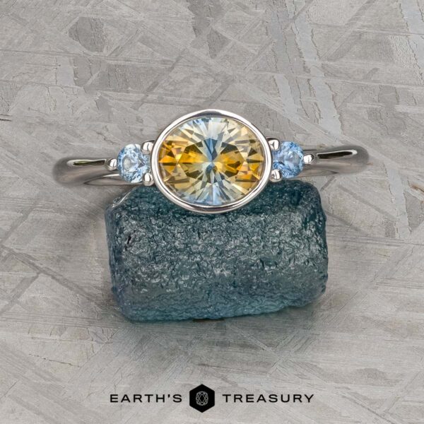 The "Evadne" ring in 14K white gold with 1.34-carat Montana sapphire The "Evadne" ring in 14K white gold, featuring a 1.34-carat round Montana sapphire between two smaller blue gems, is shown on a rough blue mineral with the "Earth's Treasury" logo below.