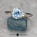 The "Deirdre" ring in platinum, featuring a 1.49-carat faceted Montana sapphire, is displayed atop a rough blue crystal with the "Earth's Treasury" branding visible below.