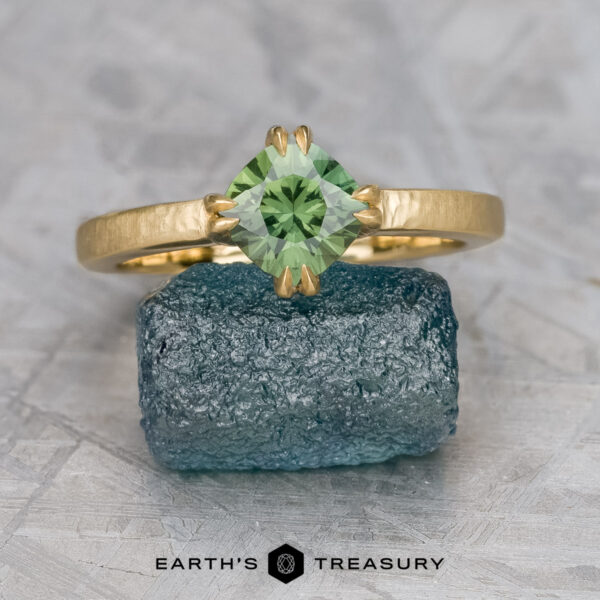 The "Capella" rign in 18K yellow gold with 1.07-carat Australian sapphire The "Capella" ring in 18K yellow gold with a 1.07-carat Australian sapphire stands on a rough blue crystal, with the "Earth's Treasury" logo and text beneath.