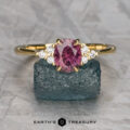The "Kacie" ring in 18K yellow gold, featuring a 1.41-carat Umba sapphire and six small white stones, is displayed on a rough greenish-blue mineral against a textured gray background.