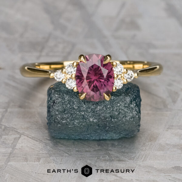The "Kacie" ring in 18K yellow gold with 1.41-carat Umba sapphire The "Kacie" ring in 18K yellow gold, featuring a 1.41-carat Umba sapphire and six small white stones, is displayed on a rough greenish-blue mineral against a textured gray background.