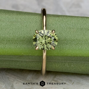 The "Begonia" ring in 14K yellow gold, featuring a 1.56-carat Australian sapphire, is displayed upright on a green stem with the Earth's Treasury logo at the bottom center.