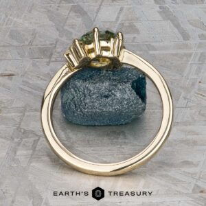 The "Nebula" ring in 14K yellow gold with a 1.54-carat Montana sapphire stands upright against a rough blue mineral on a textured gray surface. The text "EARTH'S TREASURY" appears at the bottom.