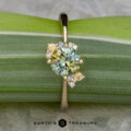 The "Nebula" ring features a 1.54-carat yellow-green Montana sapphire in 14K yellow gold, accented with small diamonds and yellow stones, beautifully set and displayed on a green striped leaf.