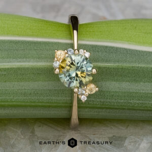 The "Nebula" ring features a 1.54-carat yellow-green Montana sapphire in 14K yellow gold, accented with small diamonds and yellow stones, beautifully set and displayed on a green striped leaf.