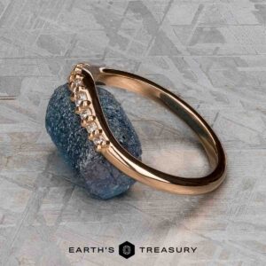 The "Hesperia" band in 20K pink gold, featuring a curved row of diamonds, is shown resting on a rough blue gemstone above the Earth’s Treasury logo on a textured gray surface.