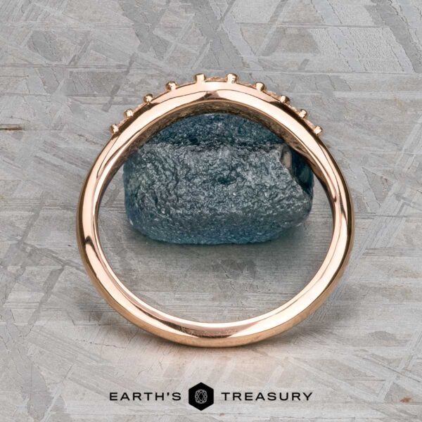 The "Hesperia" band in 20K pink gold The "Hesperia" band in 20K pink gold stands upright, framing a rough blue gemstone on a textured gray surface, with the "Earth's Treasury" logo at the bottom.