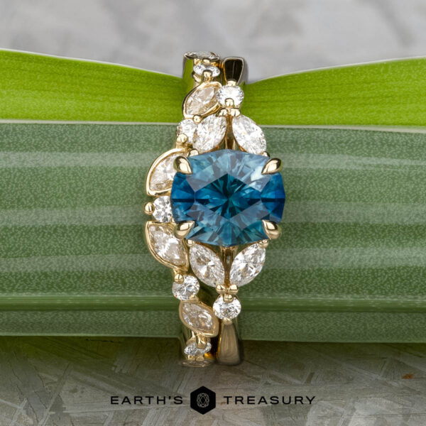 The "Cattleya" ring in 14K yellow gold with 2.22-carat Montana sapphire, alongside the "Thames" band in 14K yellow gold The "Cattleya" ring in 14K yellow gold with a 2.22-carat Montana sapphire is shown upright beside the "Thames" band, both set in gold against green leaves.