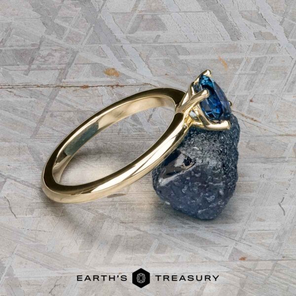 The "Clarice" ring in 14K yellow gold with 0.97-carat Montana sapphire The "Clarice" ring in 14K yellow gold with a 0.97-carat Montana sapphire stands upright, balanced on a rough blue stone, with the "Earth's Treasury" logo at the bottom.