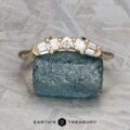 The "Mandevilla" band in 14K yellow gold, featuring three round and four baguette diamonds, is displayed on a rough blue stone with the "Earth's Treasury" logo and text below.