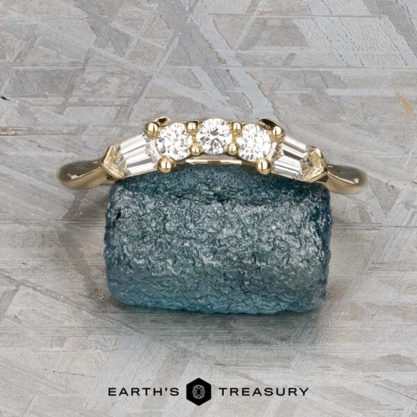 The "Mandevilla" band in 14K yellow gold The "Mandevilla" band in 14K yellow gold, featuring three round and four baguette diamonds, is displayed on a rough blue stone with the "Earth's Treasury" logo and text below.