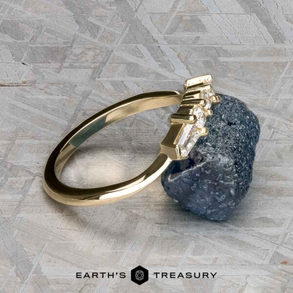 The "Mandevilla" band in 14K yellow gold The "Mandevilla" band in 14K yellow gold, featuring a row of diamonds, rests against a rough, dark stone on a textured gray background. The "Earth’s Treasury" logo appears at the bottom of the image.