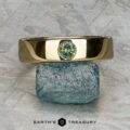 The "Logan" sapphire ring in 18K yellow gold, featuring a round green gemstone, is balanced on a rough blue-green stone atop a textured gray surface with the "Earth's Treasury" logo below.