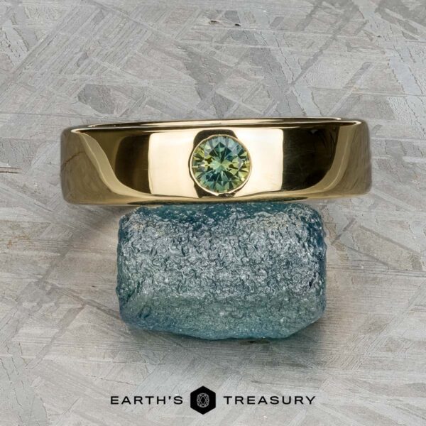 The "Logan" sapphire ring in 18K yellow gold The "Logan" sapphire ring in 18K yellow gold, featuring a round green gemstone, is balanced on a rough blue-green stone atop a textured gray surface with the "Earth's Treasury" logo below.
