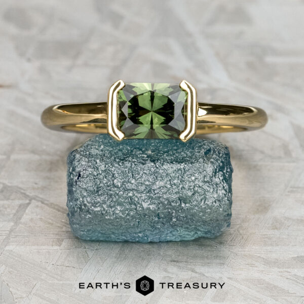 The "Larisa" ring in 18K yellow gold with 1.04-carat Australian sapphire The "Larisa" ring in 18K yellow gold featuring a 1.04-carat Australian sapphire is displayed on a rough blue stone, with the "Earth’s Treasury" logo and name at the bottom.