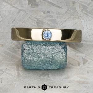 The "Denali" ring in 14K yellow gold, featuring a round blue gemstone, is balanced on a rough rectangular blue stone. The backdrop is a textured, light surface with "EARTH'S TREASURY" displayed at the bottom.