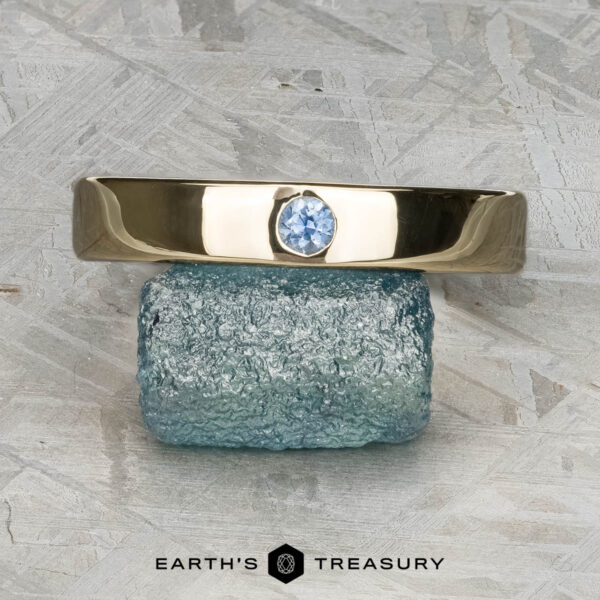 The "Denali" ring in 14K yellow gold The "Denali" ring in 14K yellow gold, featuring a round blue gemstone, is balanced on a rough rectangular blue stone. The backdrop is a textured, light surface with "EARTH'S TREASURY" displayed at the bottom.