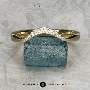 The "Rhine" band in 14K yellow gold, featuring a curved row of small white gemstones, rests on a rough blue mineral. A light, textured background appears behind with "EARTH'S TREASURY" written at the bottom.