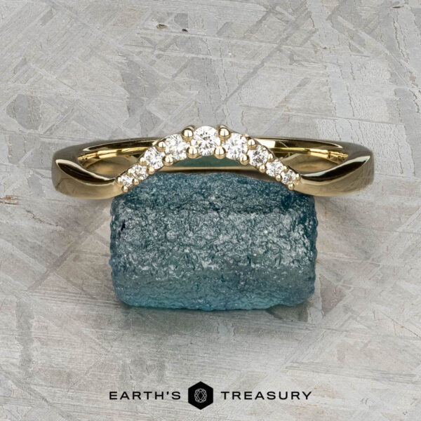 The "Rhine" band in 14K yellow gold The "Rhine" band in 14K yellow gold, featuring a curved row of small white gemstones, rests on a rough blue mineral. A light, textured background appears behind with "EARTH'S TREASURY" written at the bottom.