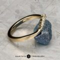The "Rhine" band in 14K yellow gold, featuring a row of small diamonds, rests against a rough blue gemstone on a textured gray surface. The text "EARTH'S TREASURY" appears at the bottom.