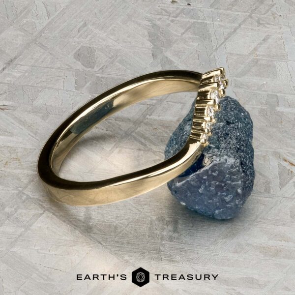 The "Rhine" band in 14K yellow gold The "Rhine" band in 14K yellow gold, featuring a row of small diamonds, rests against a rough blue gemstone on a textured gray surface. The text "EARTH'S TREASURY" appears at the bottom.