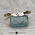 The Minimal “Lonicera” band in 14K rose gold is elegantly balanced on a rough blue gemstone, with “EARTH’S TREASURY” displayed beneath on a textured gray background.