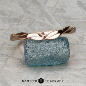 The Minimal “Lonicera” band in 14K rose gold is elegantly balanced on a rough blue gemstone, with “EARTH’S TREASURY” displayed beneath on a textured gray background.