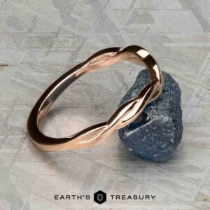 The Minimal “Lonicera” band in 14K rose gold is elegantly balanced on a rough blue gemstone, with “EARTH’S TREASURY” displayed beneath on a textured gray background.