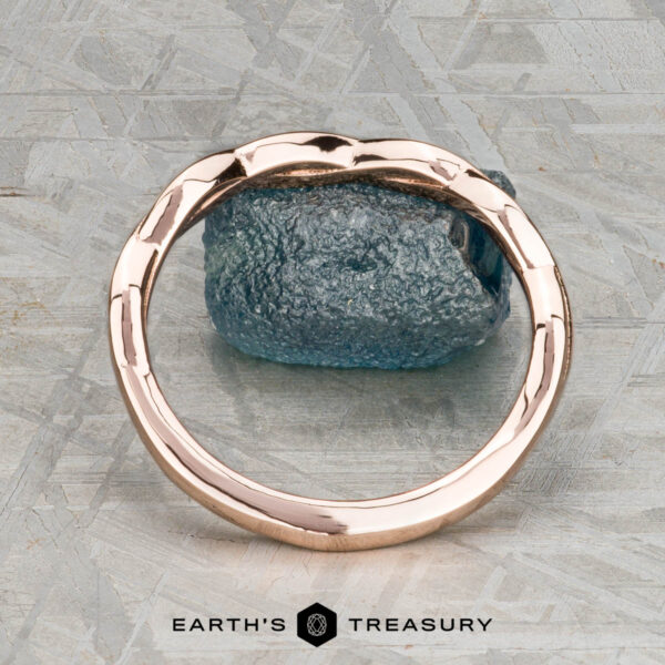 The Minimal "Lonicera" band in 14K rose gold The Minimal “Lonicera” band in 14K rose gold is elegantly balanced on a rough blue gemstone, with “EARTH’S TREASURY” displayed beneath on a textured gray background.