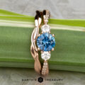 The "Lonicera" ring in 14K rose gold features a 0.93-carat Montana sapphire center, two side diamonds, and small band diamonds, shown with the minimal "Lonicera" band in 14K rose gold against a green stem.