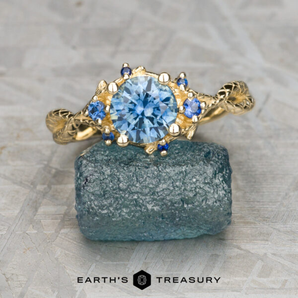 The "Juniper" ring in 14K yellow gold with 1.07-carat Montana sapphire The "Juniper" ring in 14K yellow gold features a 1.07-carat round Montana sapphire centerpiece and smaller blue gems, displayed on a blue-green mineral with "Earth's Treasury" below.