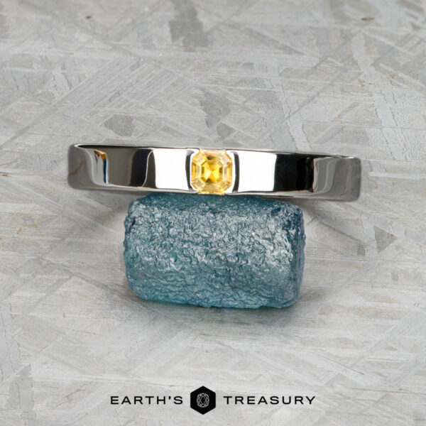 The "Kilimanjaro" ring in 14K white gold The "Kilimanjaro" ring in 14K white gold, featuring a rectangular yellow gemstone, is displayed balanced on a rough blue stone with the Earth's Treasury logo beneath.