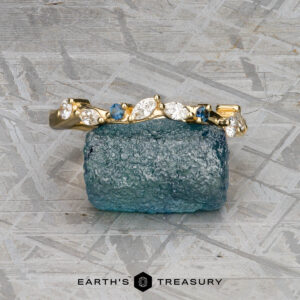 The "Posey" band in 18K yellow gold with a geometric design is displayed upright before a rough blue gemstone, set against a textured gray background with the Earth's Treasury logo below.
