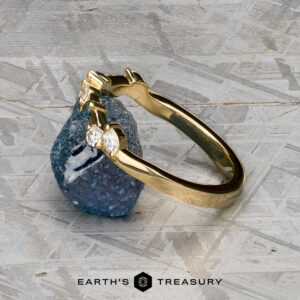 The "Posey" band in 18K yellow gold with a geometric design is displayed upright before a rough blue gemstone, set against a textured gray background with the Earth's Treasury logo below.