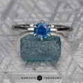 The "Anemone" ring in 14K white gold with a 0.83-carat Montana sapphire is displayed on a rough blue crystal atop a patterned metallic surface featuring the “EARTH’S TREASURY” logo.