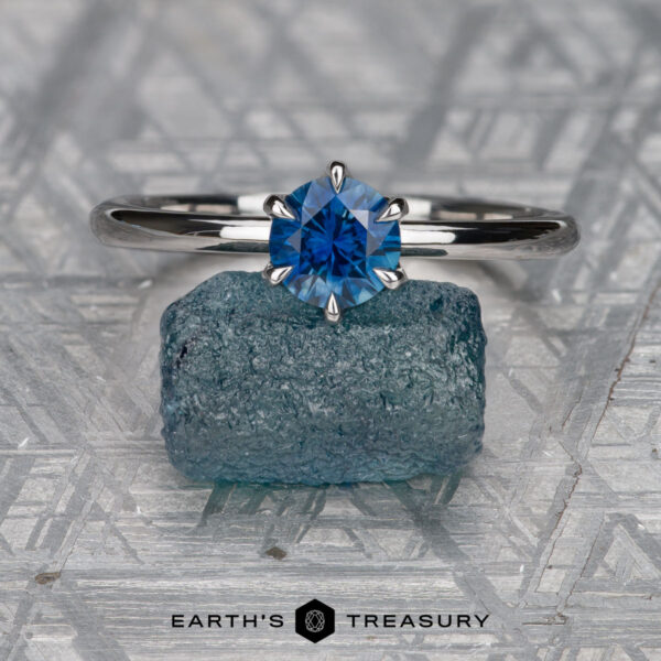 The "Anemone" ring in 14K white gold with 0.83-carat Montana sapphire The "Anemone" ring in 14K white gold with a 0.83-carat Montana sapphire is displayed on a rough blue crystal atop a patterned metallic surface featuring the “EARTH’S TREASURY” logo.