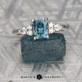 The "Vela" ring in 14K white gold features a 0.93-carat rectangular Montana sapphire and small round diamonds, displayed on a rough blue stone with the text "EARTH'S TREASURY" beneath it.
