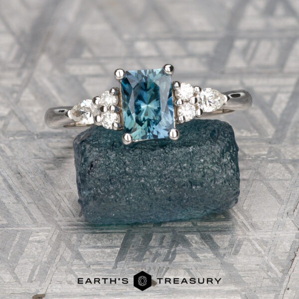The "Vela" ring in 14K white gold with 0.93-carat Montana sapphire The "Vela" ring in 14K white gold features a 0.93-carat rectangular Montana sapphire and small round diamonds, displayed on a rough blue stone with the text "EARTH'S TREASURY" beneath it.
