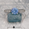 The "Aquila" ring in 14K white gold features a 1.55-carat Ceylon sapphire and accent stones on the band, displayed on a rough blue crystal with a geometric background and "EARTH'S TREASURY" text below.