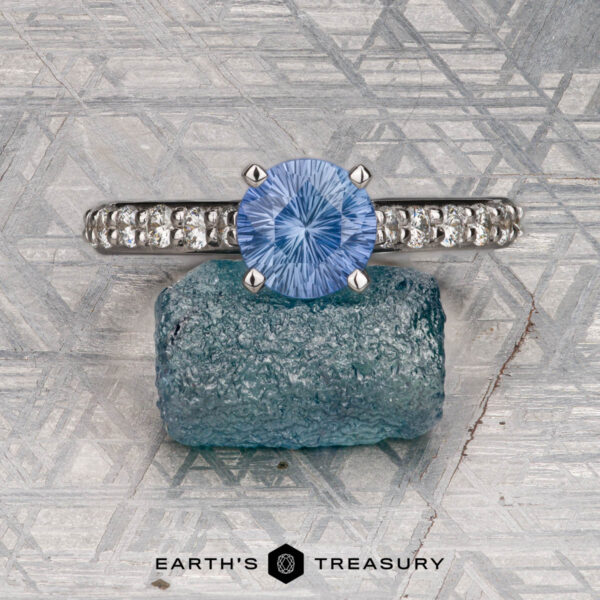 The "Aquila" ring in 14K white gold with 1.55-carat Ceylon sapphire The "Aquila" ring in 14K white gold features a 1.55-carat Ceylon sapphire and accent stones on the band, displayed on a rough blue crystal with a geometric background and "EARTH'S TREASURY" text below.