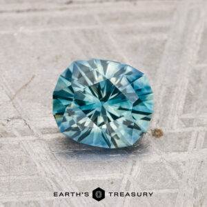 A 1.17-carat heated Montana Sapphire, round and brilliant-cut with blue-green hues, rests on a textured gray surface with "EARTH'S TREASURY" displayed below.