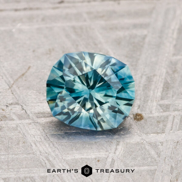 1.17-Carat Montana Sapphire (Heated) A 1.17-carat heated Montana Sapphire, round and brilliant-cut with blue-green hues, rests on a textured gray surface with "EARTH'S TREASURY" displayed below.