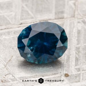 A 1.94-carat heated Montana sapphire is showcased on a light, textured surface with "EARTH'S TREASURY" displayed at the bottom.